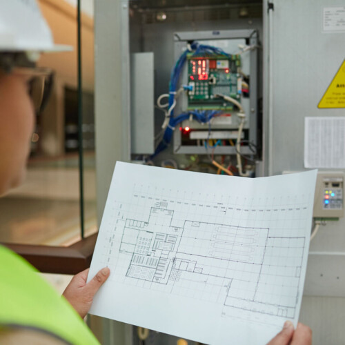 Female worker holding blueprints while inspecting electrical switchboard at construction site, copy space